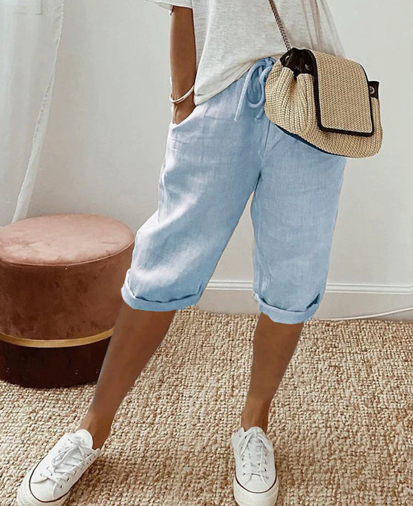 Vrouw in casual outfit met lichtblauwe linnen shorts, witte sneakers en een beige schoudertas. Perfecte zomermode, comfortabele en stijlvolle look.