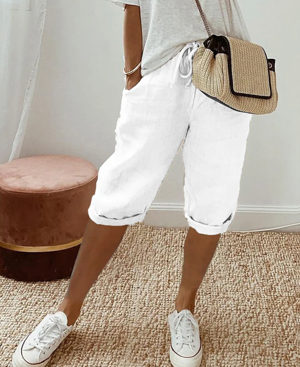 Vrouw in casual outfit met witte linnen bermuda shorts, beige schoudertas en witte sneakers op een beige tapijt. Perfect voor zomermode en comfort.