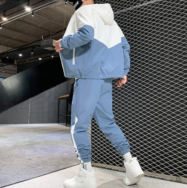Man in blauw-wit trainingspak met capuchon en witte sneakers, moderne sportieve kleding, industriële achtergrond, streetwear mode.