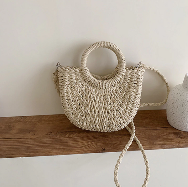 Rieten handtas met ronde handgrepen en schouderriem, rustend op houten plank. Stijlvolle zomertas, perfect voor stranduitjes en casual outfits.
