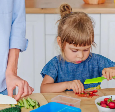 Kleuter in blauwe jurk snijdt groenten met kindvriendelijk mes in keuken. Gezonde voeding, koken met kinderen, educatieve activiteiten.