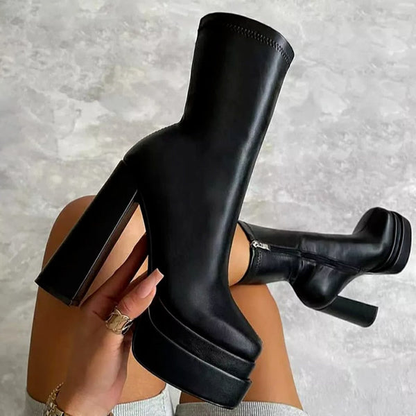 Zwarte leren platformlaarzen met hoge hakken, gedragen door een persoon zittend op een grijze vloer. Trendy damesmode, herfst/winter schoenen.