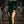 Vrouw in elegante groene fluwelen avondjurk met hoge split, staand op trap, met clutch en opvallende oorbellen. Perfect voor galafeest of avondje uit.