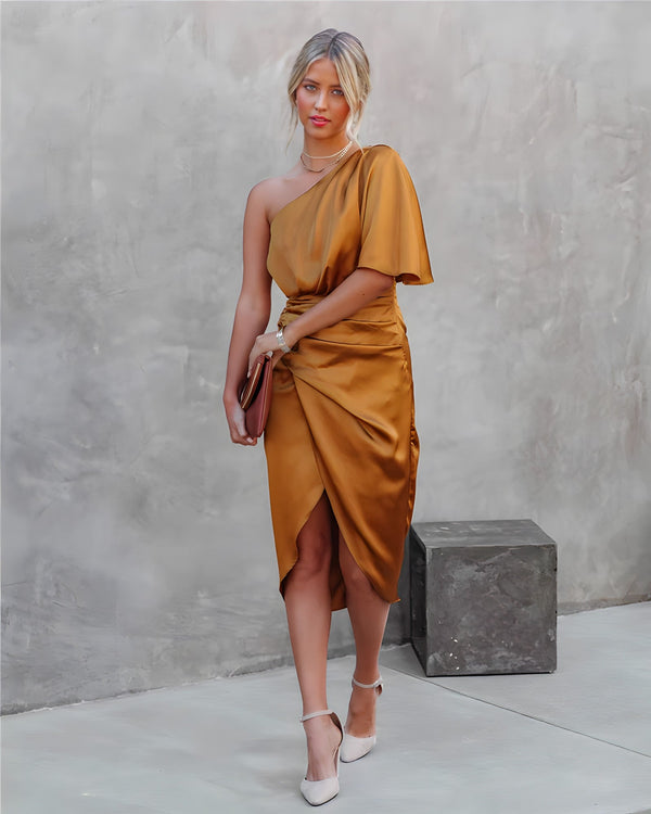 Vrouw in elegante, goudkleurige asymmetrische jurk met clutch en hakken, poseert tegen een minimalistische betonnen achtergrond. Fashion, avondkleding.