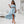 Vrouw in lichtblauwe off-shoulder jurk met ruches, hoge split en sandalen, poseert in moderne woonkamer. Trendy zomeroutfit, elegante mode.