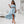 Vrouw in lichtblauwe off-shoulder jurk met ruches, hoge split en sandalen, poseert in moderne witte woonkamer. Zomerjurk, mode, elegante outfit.