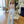 Elegante vrouw in stijlvolle outfit met doorschijnende top en wijde broek, staand in een luxe woonkamer met moderne inrichting en grote planten.