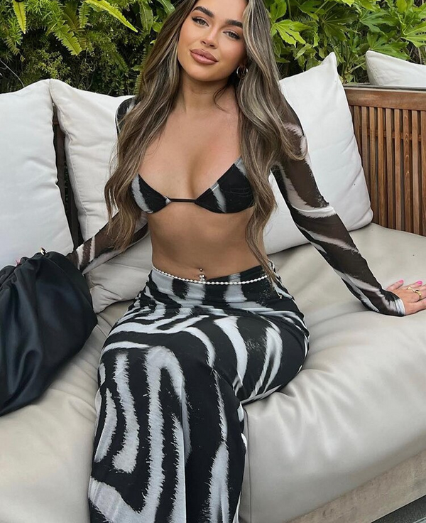 Vrouw in zwart-witte zebraprint outfit, zittend op een beige bank met groene planten op de achtergrond. Trendy mode, zomerstijl, buitenlounge.
