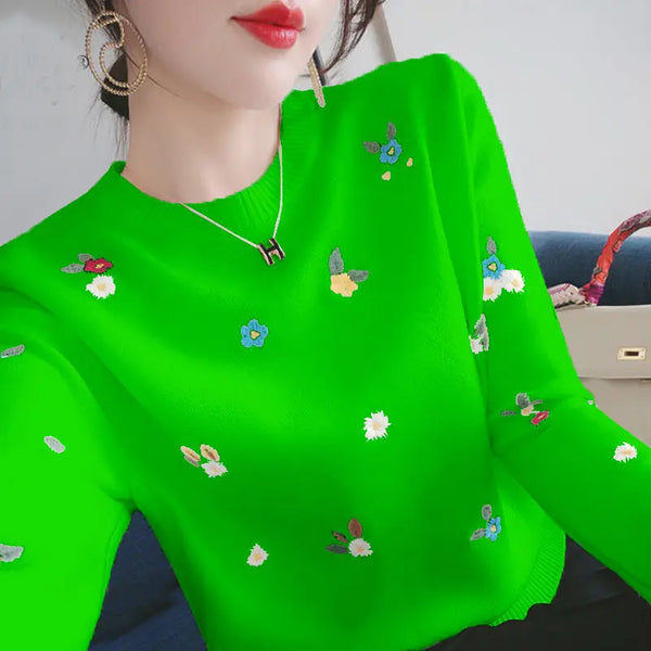 Vrouw in felgroene trui met kleurrijke bloemenborduursels, gouden oorbellen en ketting, zittend op blauwe bank. Modieuze dameskleding, trendy stijl.