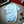 Lichtblauw heren T-shirt met korte mouwen, houten knoopdetail, op grijze vloer naast theepot en plant. Casual zomeroutfit.