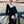 Vrouw in elegante zwarte jurk met witte ruches en knopen, poseert op brug met uitzicht op rivier en historische gebouwen in Boedapest bij zonsondergang.