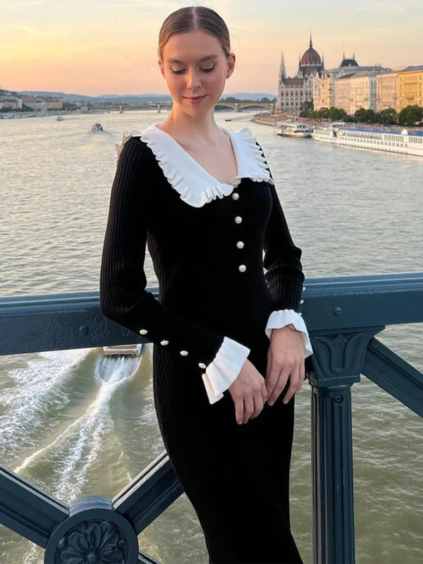 Vrouw in elegante zwarte jurk met witte ruches en knopen, poseert op brug met uitzicht op rivier en historische gebouwen in Boedapest bij zonsondergang.
