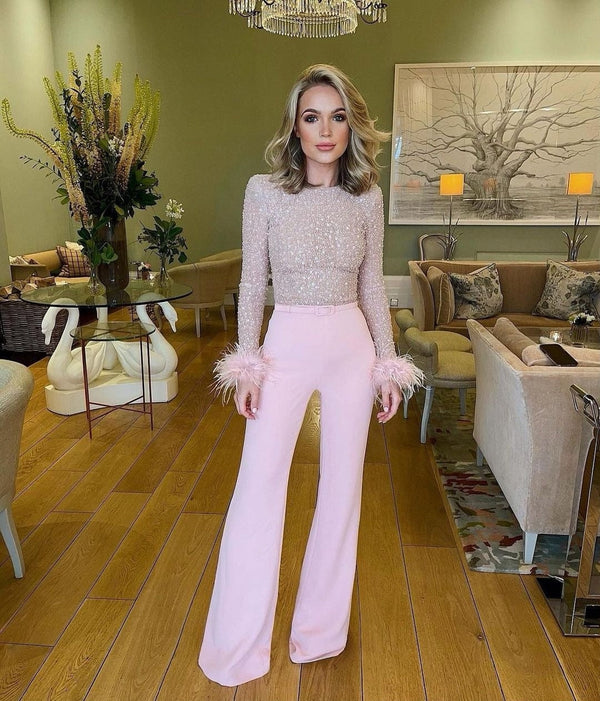 Vrouw in elegante roze broek en glinsterende top met veren, staand in stijlvolle woonkamer met groene muren en moderne decoratie. Luxe mode, interieur.