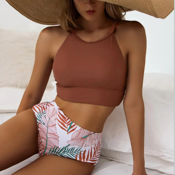 Bruin crop top en tropische bikini met bladerenprint, gedragen door vrouw met grote zonnehoed. Zomerse strandmode, trendy badkleding.