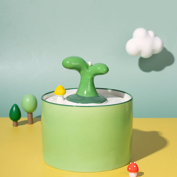 Groene keramische kattenfontein met plantmotief, omgeven door miniatuur paddenstoelen en bomen, tegen een pastelgroene achtergrond met wolk.