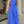 Vrouw in blauwe jumpsuit met abstracte zwarte lijnen, halternek en wijde pijpen, houdt groene clutch vast. Stijlvolle mode voor dames.