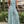 Vrouw in lichtblauwe, mouwloze jumpsuit met abstract patroon, staand buiten bij groene planten. Trendy damesmode, zomeroutfit, casual stijl.