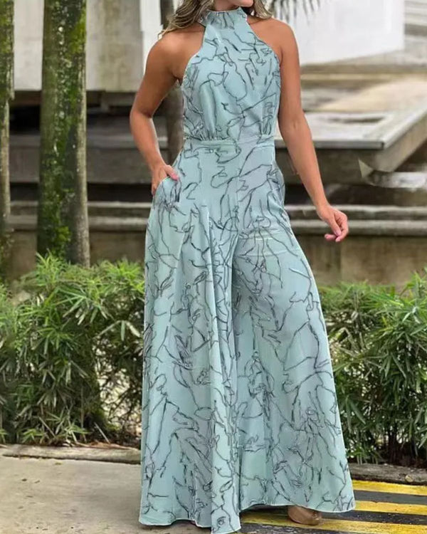 Vrouw in lichtblauwe, mouwloze jumpsuit met abstract patroon, staand buiten bij groene planten. Trendy damesmode, zomeroutfit, casual stijl.