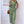 Vrouw in groene jumpsuit met knoopsluiting en strikceintuur, draagt bruine schoudertas. Stijlvolle zomermode tegen witte bakstenen muur.