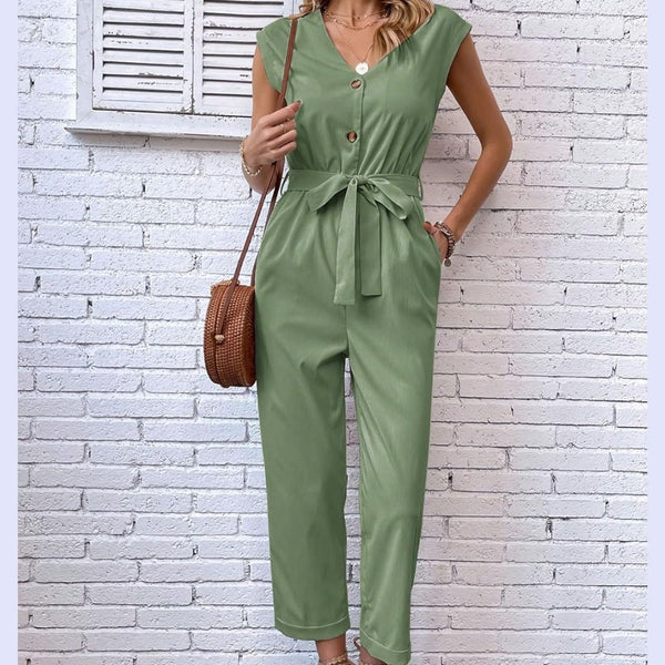 Vrouw in groene jumpsuit met knoopsluiting en strikceintuur, draagt bruine schoudertas. Stijlvolle zomermode tegen witte bakstenen muur.