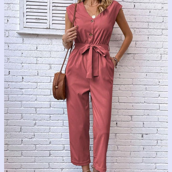 Vrouw in roze jumpsuit met knoopsluiting en strikceintuur, draagt bruine schoudertas. Stijlvolle damesmode tegen witte bakstenen muur.