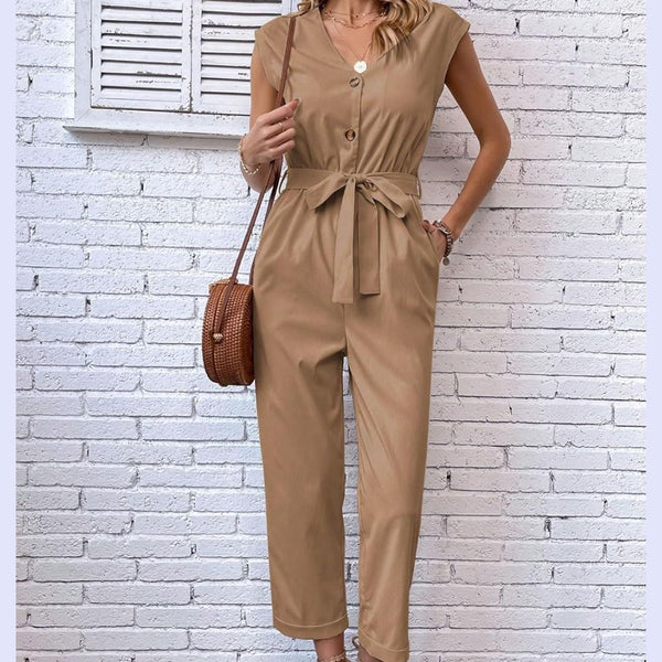 Vrouw in stijlvolle beige jumpsuit met knoopsluiting en strikceintuur, poseert voor witte bakstenen muur. Trendy mode, casual chic outfit.