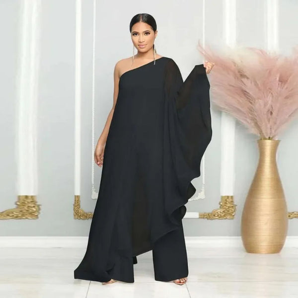 Elegante vrouw in zwarte asymmetrische avondjurk met lange sluier, staand voor een decoratieve muur met gouden accenten en vaas met pampasgras.
