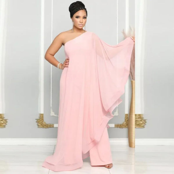 Vrouw in elegante roze asymmetrische avondjurk met wijde mouw, staand voor een grijze muur met gouden accenten. Perfect voor galafeest of bruiloft.