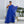 Elegante vrouw in blauwe asymmetrische avondjurk met transparante cape, staand voor een moderne grijze muur met planten. Perfect voor galafeest.