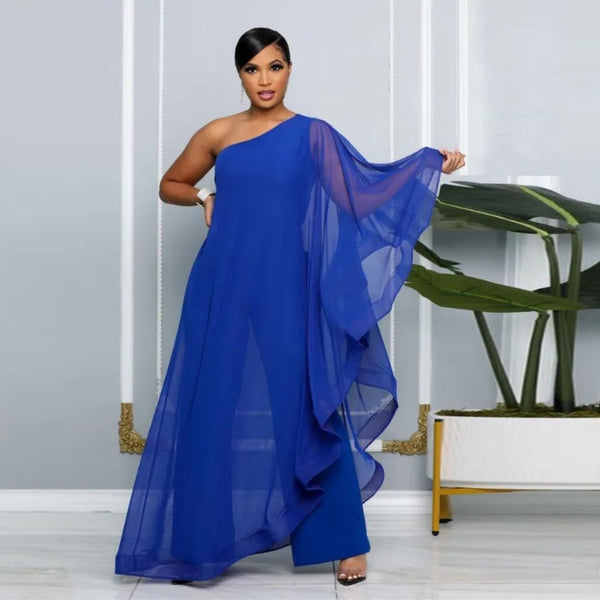 Elegante vrouw in blauwe asymmetrische avondjurk met transparante cape, staand voor een moderne grijze muur met planten. Perfect voor galafeest.