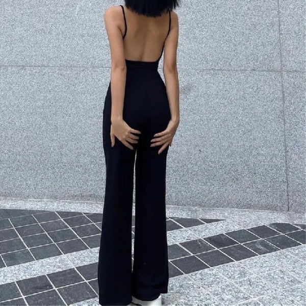 Vrouw in zwarte, rugloze jumpsuit met wijde pijpen, staand op betegelde vloer voor grijze muur. Trendy mode, minimalistisch design, elegante outfit.