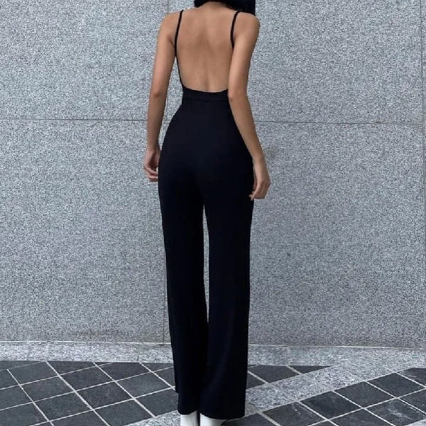 Vrouw in elegante zwarte jumpsuit met open rug, staand tegen grijze stenen muur. Modieuze dameskleding, minimalistisch design.