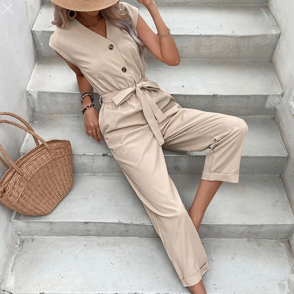 Vrouw in beige jumpsuit met knopen en strikceintuur, zittend op betonnen trap. Strohoed en rieten tas naast haar. Zomermode, casual outfit.