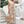 Vrouw in beige jumpsuit met knopen en strikceintuur, houdt een gevlochten rieten tas vast. Stijlvolle zomermode, casual outfit, trendy kleding.
