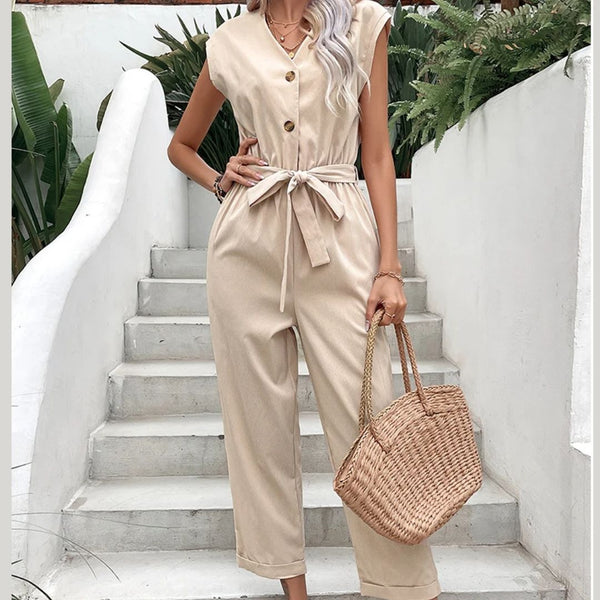 Vrouw in beige jumpsuit met knoopsluiting en strikceintuur, draagt een gevlochten rieten tas. Stijlvolle zomermode op witte trap met groene planten.