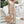 Vrouw in beige jumpsuit met knoopsluiting en strikceintuur, draagt een gevlochten rieten tas. Stijlvolle zomermode, casual outfit, trap op achtergrond.