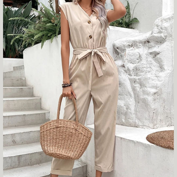 Vrouw in beige jumpsuit met knoopsluiting en strikceintuur, draagt een gevlochten rieten tas. Stijlvolle zomermode, casual outfit, trap op achtergrond.