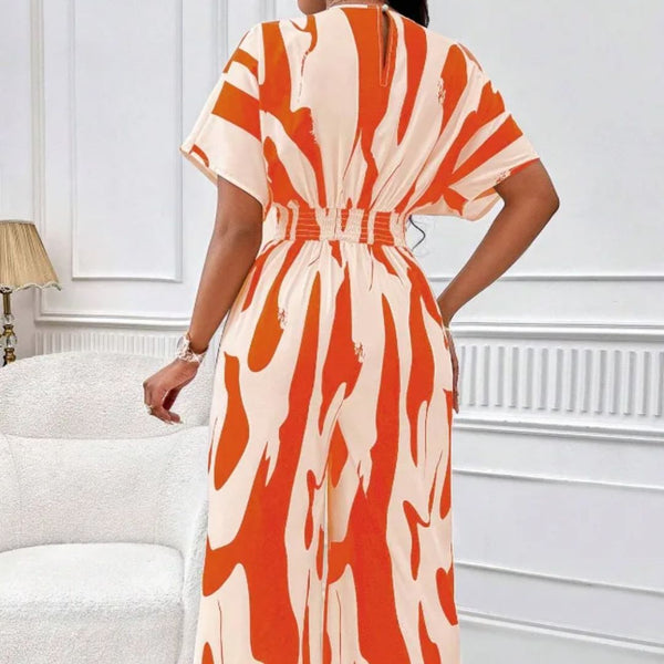 Vrouw in oranje-witte maxi-jurk met abstract patroon, staand in moderne woonkamer. Trendy damesmode, zomerjurk, elegant design.