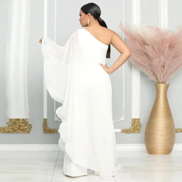 Vrouw in elegante witte asymmetrische avondjurk met één schouder, staand voor een grijze muur met gouden accenten en een vaas met pampasgras.