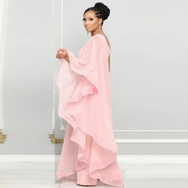Vrouw in elegante roze avondjurk met lange mouwen en golvende lagen, staand tegen een grijze achtergrond. Perfect voor mode en avondkleding.