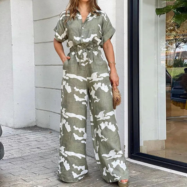 Vrouw in groene camouflage jumpsuit met korte mouwen, staand buiten bij glazen deur. Modieuze dameskleding, casual zomeroutfit.