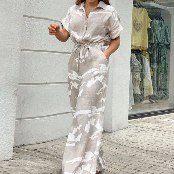 Vrouw in beige-witte linnen jumpsuit met camouflageprint, wandelend op stoep voor winkel. Modieuze dameskleding, casual zomeroutfit.