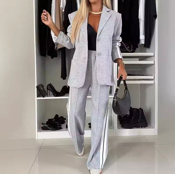 Grijze damespak met witte strepen, gedragen door vrouw in kledingkast. Modieuze zakelijke outfit, inclusief blazer en broek, met zwarte top en tas.
