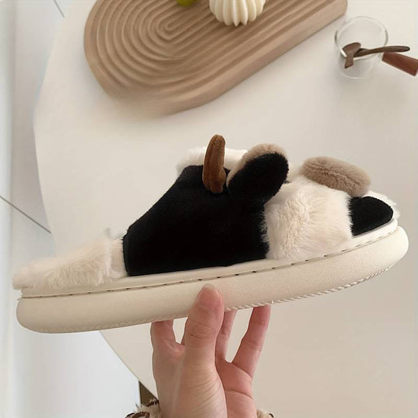 Hand houdt een pluche pantoffel vast met zwart-wit koeienprint en bruine details, op een witte achtergrond. Comfortabele, modieuze huisslippers.