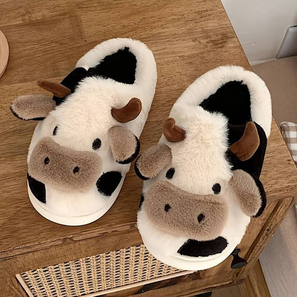 Pluche pantoffels in de vorm van een koe, wit en zwart met bruine hoorns, op houten kast. Perfect voor warme voeten en schattig design.