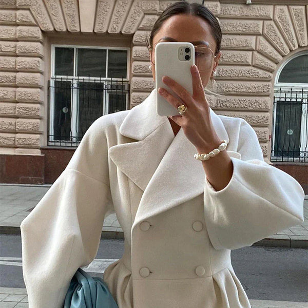 Vrouw in stijlvolle witte wollen jas met dubbele rij knopen, maakt selfie buiten. Trendy mode, herfstkleding, stedelijke achtergrond.