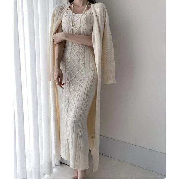 Vrouw in elegante witte gebreide jurk met kabelpatroon, lange mouwen en open voorzijde, staand bij een raam. Perfect voor stijlvolle wintermode.