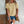 Vrouw in gele geperforeerde top en denim shorts, met bruine schoudertas en koffiebeker, tegen witte bakstenen muur. Zomermode, casual outfit.