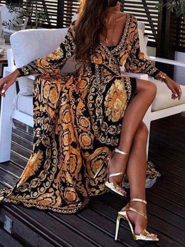 Vrouw in elegante, lange jurk met barokprint en gouden sandalen, zittend op witte stoel op houten terras. Luxe mode, zomerjurk, stijlvolle outfit.