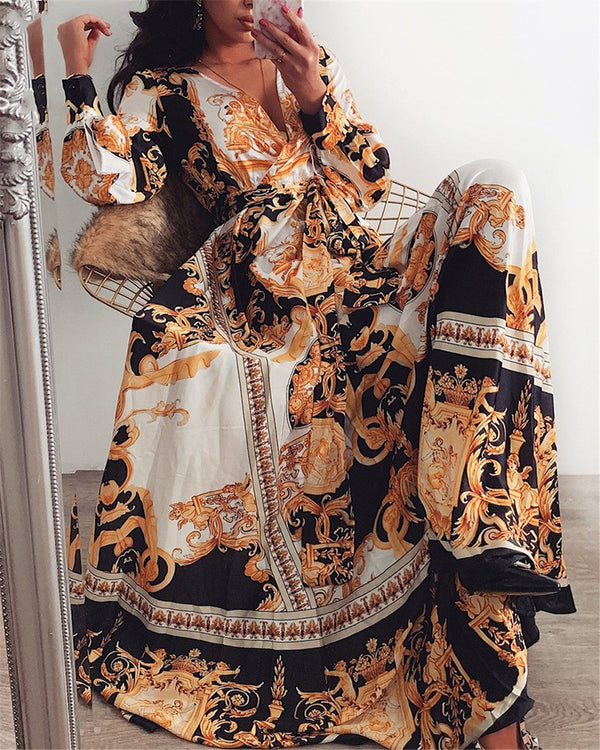 Vrouw in elegante, lange jurk met barokprint, zwart-goud-wit, zittend op stoel. Luxe mode, stijlvolle avondkleding, trendy design.